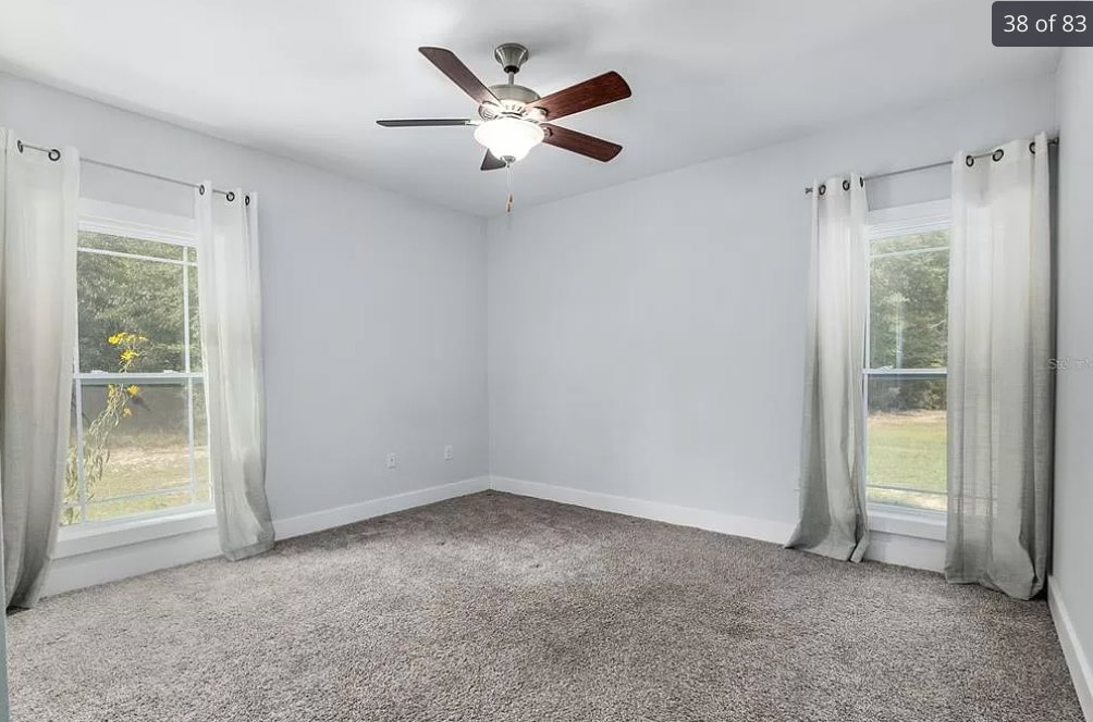 Ceiling fan with light fixture above carpeted floor, window with white curtains overlooking grass and yellow flower, plaster walls