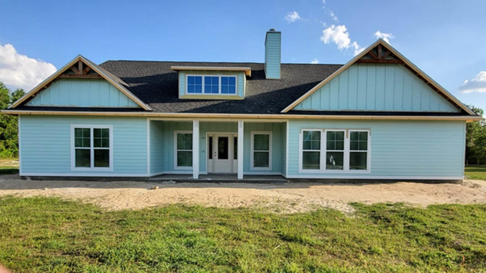 Blue siding house with white trim, front porch, white door with glass window, manicured green lawn, large front window reflecting sky and clouds.