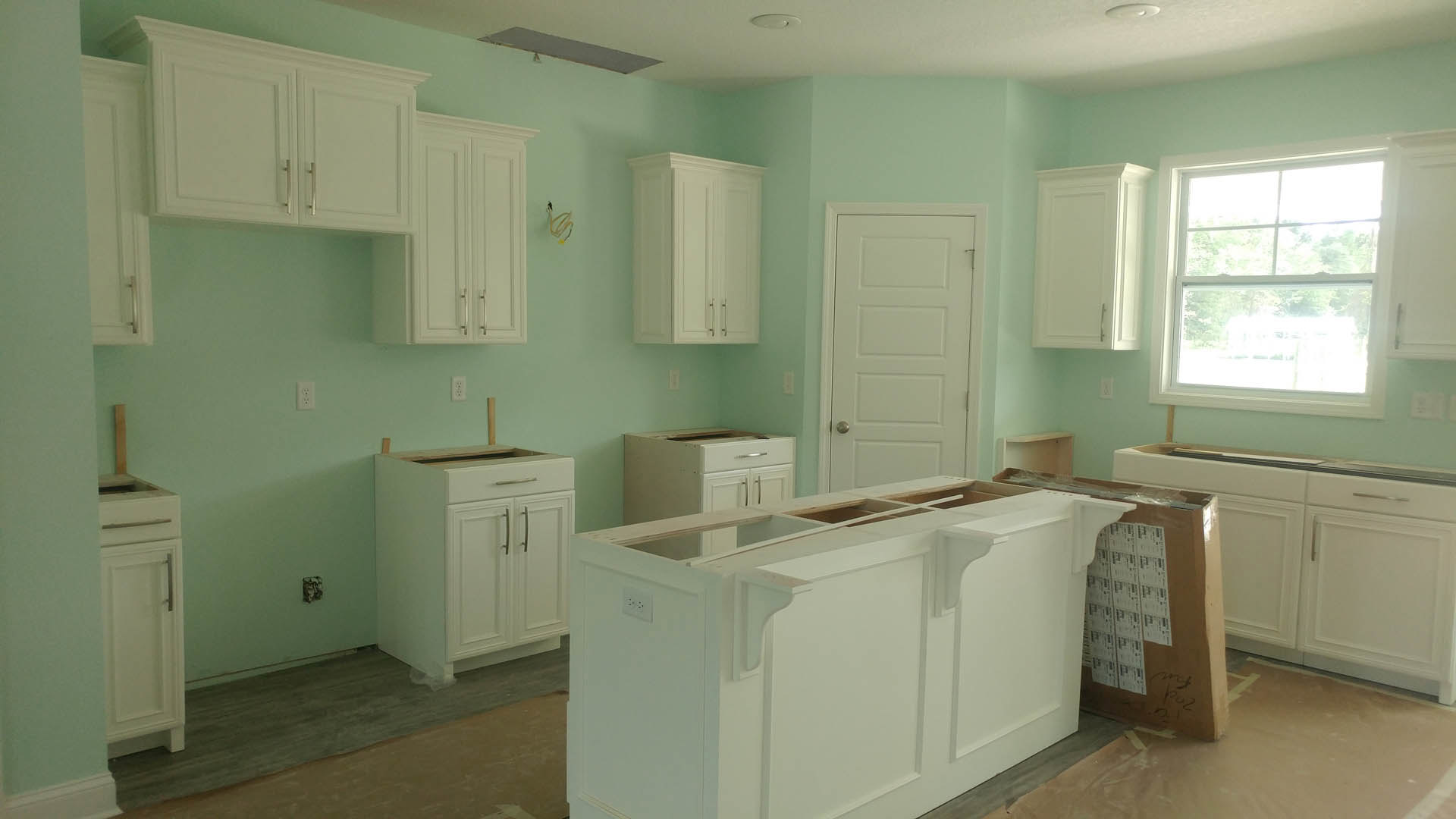 White kitchen cabinets with silver handles, green accent wall, window overlooking trees, white door, light-colored countertop, wood flooring