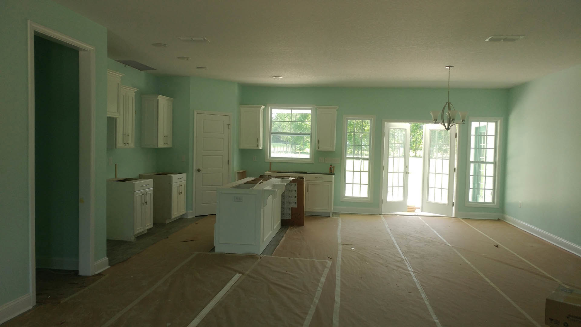 White kitchen cabinets, light blue painted walls, white door with silver handle, cardboard sheet on light flooring, window with white frame showing green trees outside