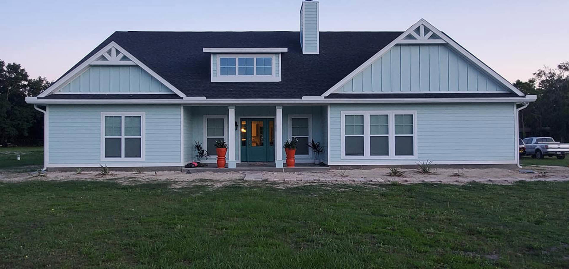 Two-story house with blue roof and blue front door, white siding, large windows, manicured green lawn, red potted plant on porch, clear sky in background