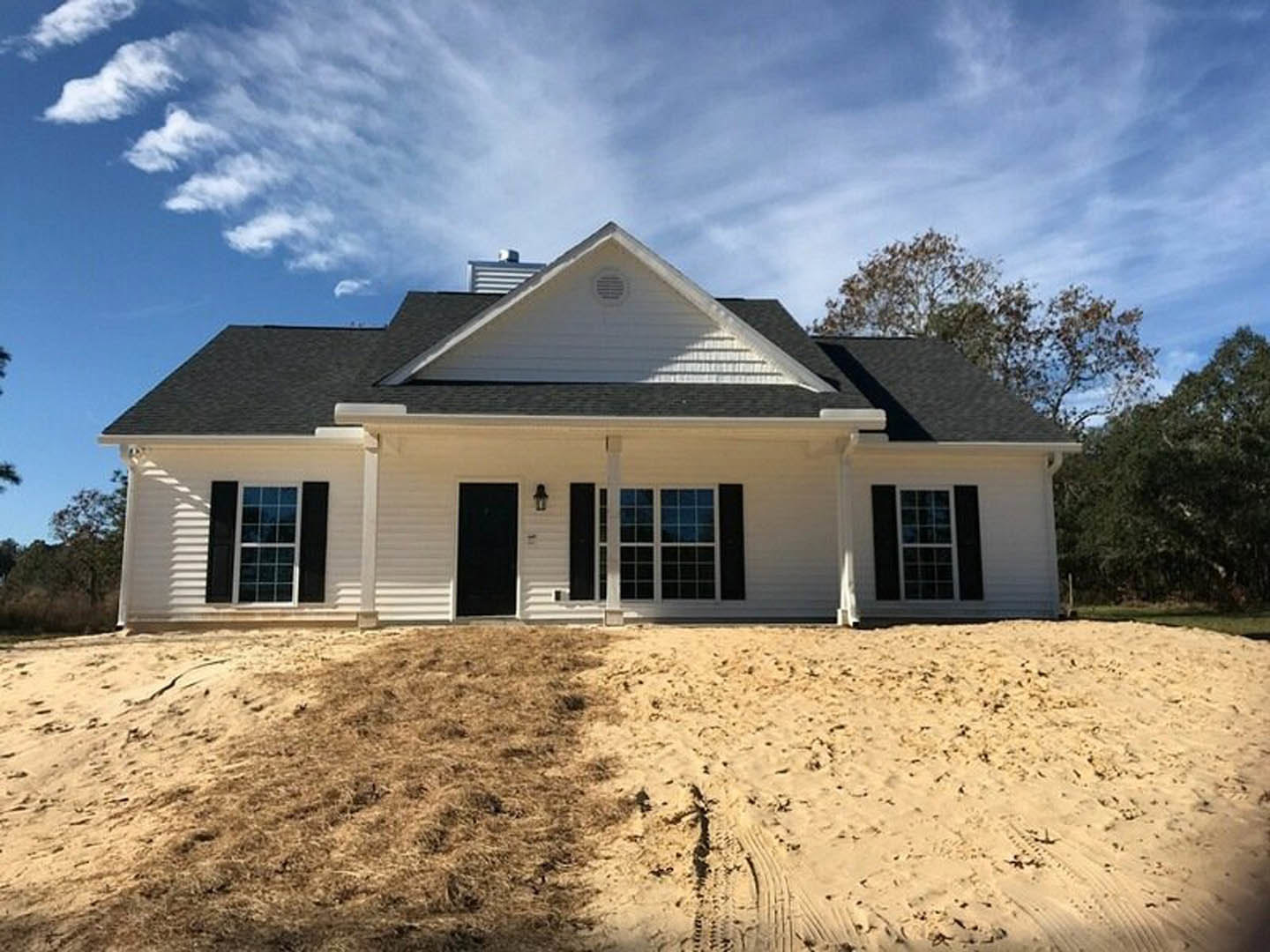 Two-story house with gray siding, white trim, and a brick chimney, surrounded by dirt and patchy grass, large windows with multiple panes, sandy area with visible footprints in