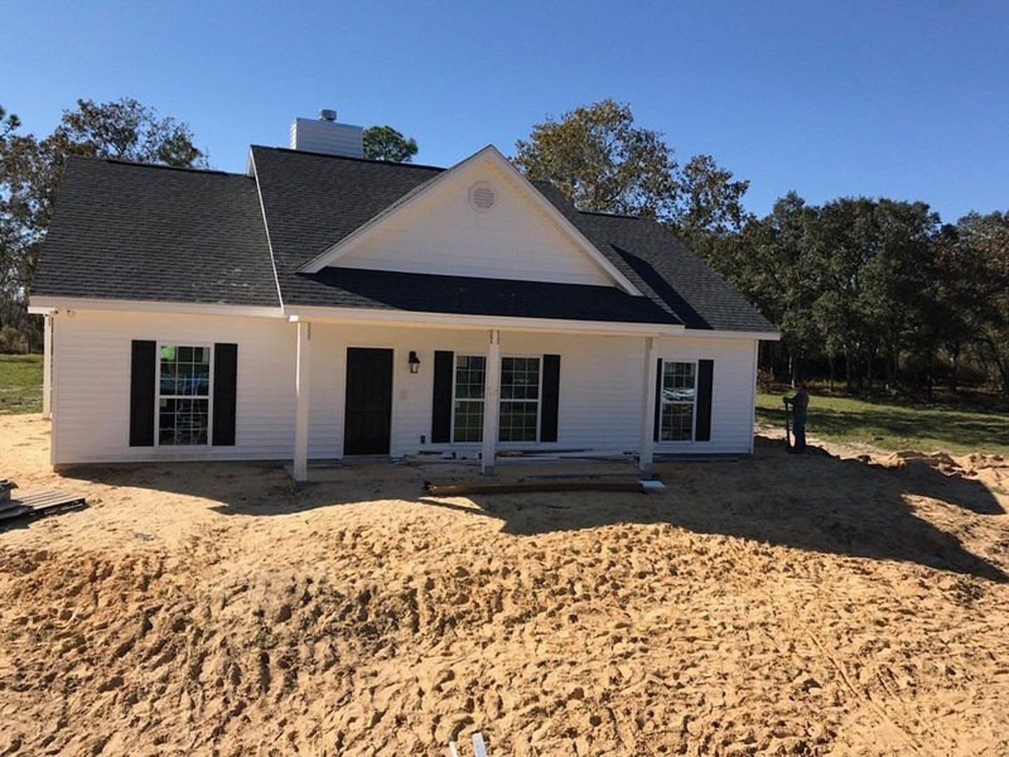 White cottage-style home with black trim, black door with white frame, pile of sand and dirt in foreground, vent on exterior wall, person holding a pole near entrance, trees and