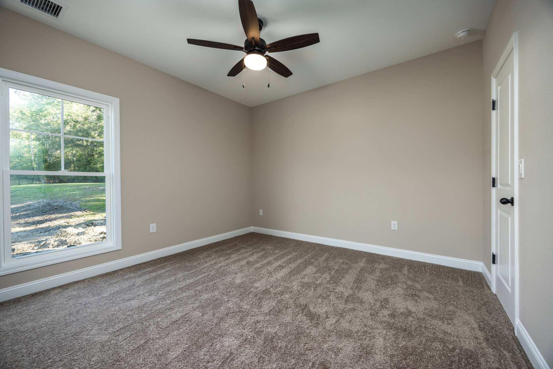 Ceiling fan with integrated light fixture mounted on white plaster ceiling, carpeted floor, large window overlooking grassy field and trees.