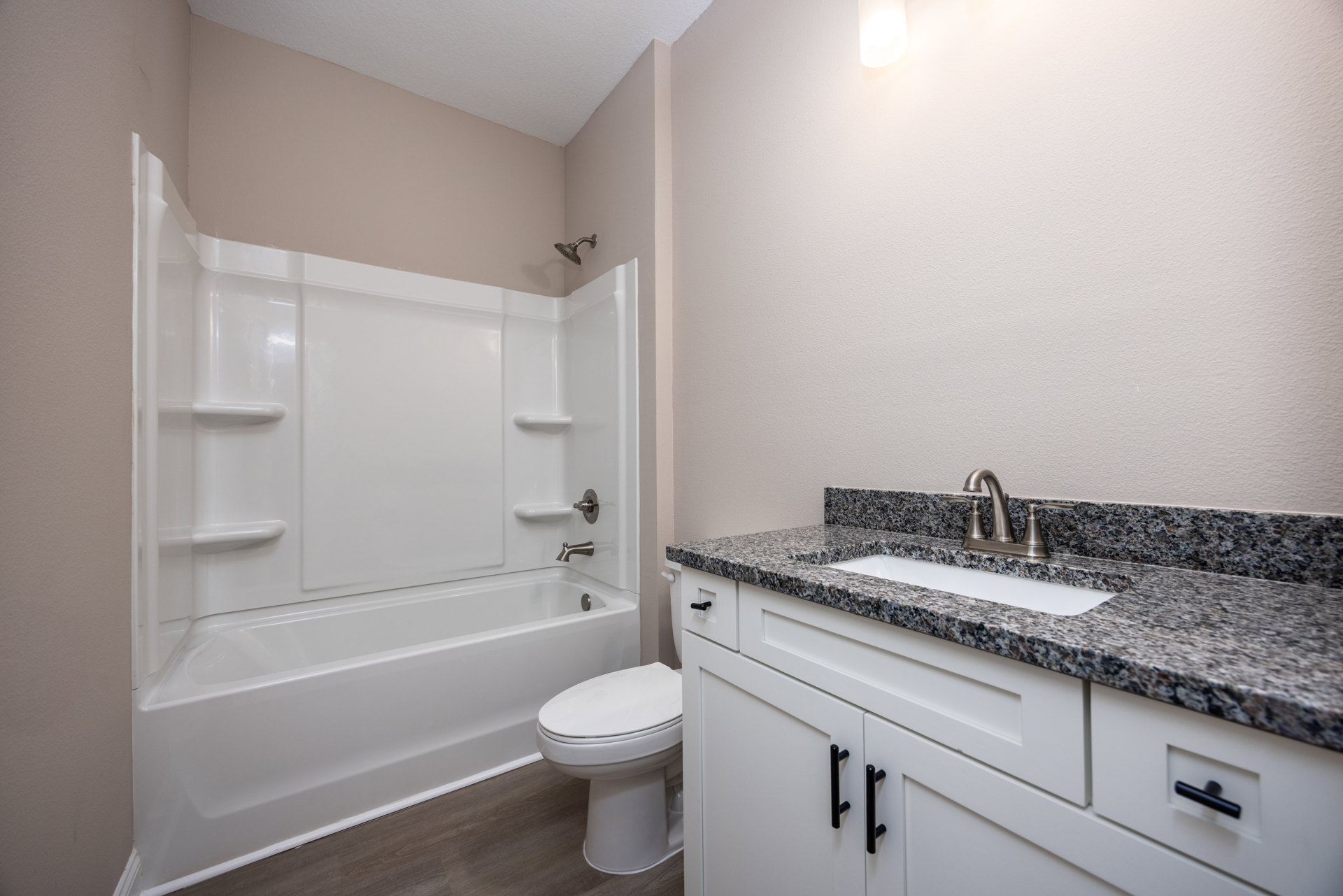 Bathroom with white bathtub, toilet with seat down, light-colored countertop, sink with chrome faucet, and cabinetry featuring black handles; neutral tile walls and floor.