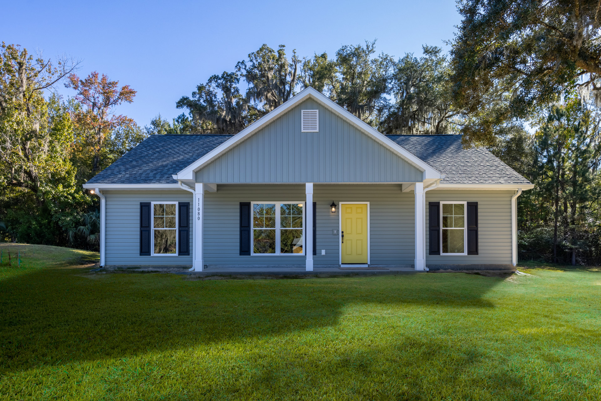 Blue house exterior with yellow front door, black handle, white vent, white-framed windows with black and blue shutters, porch, grass lawn, and small white house visible in the
