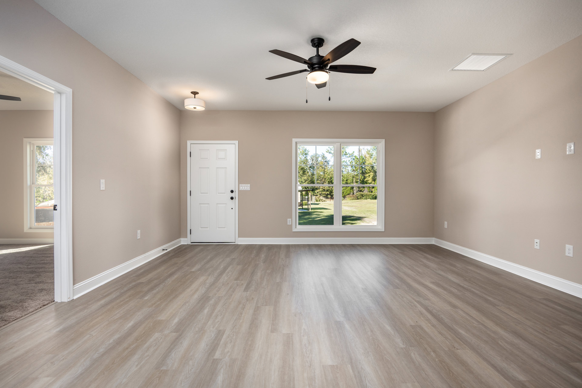 Ceiling fan with light fixture above wood flooring, white trim, white door with black knobs, window overlooking yard and trees, plaster walls and ceiling