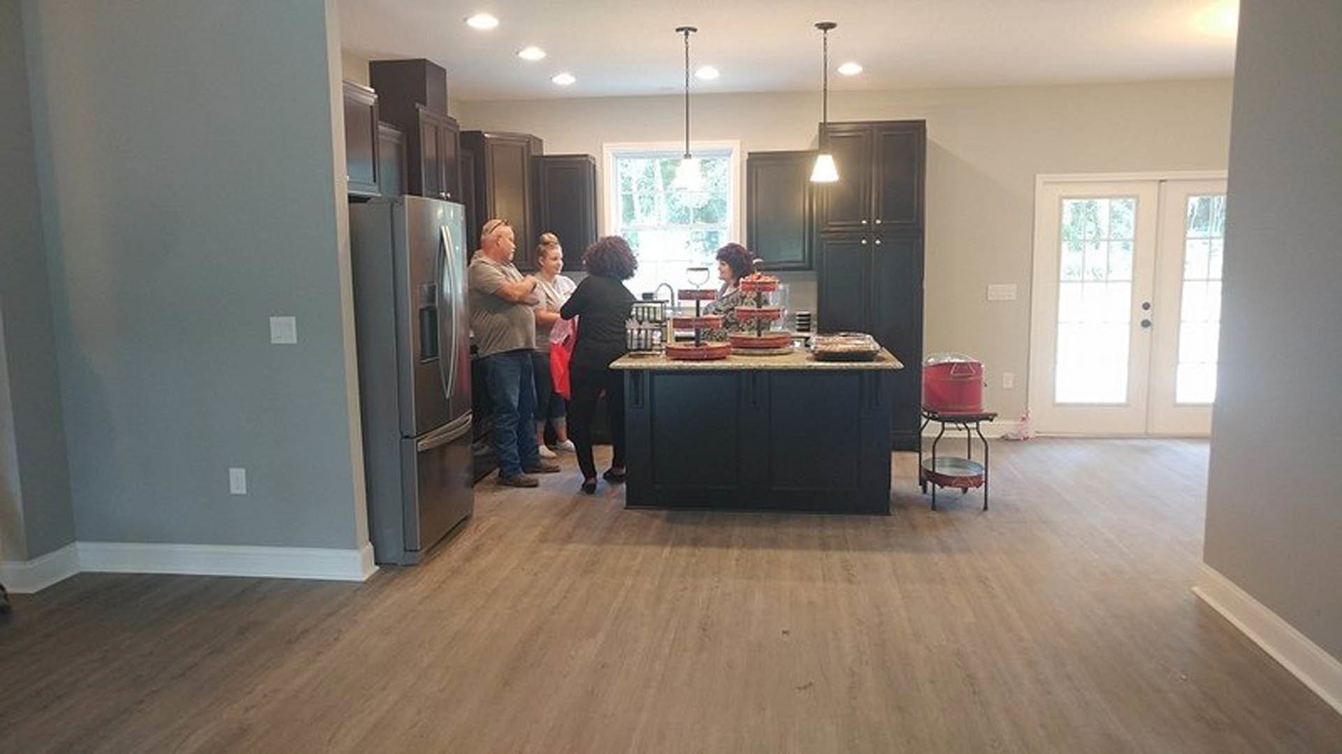Quartz kitchen island surrounded by people, black cabinetry, stainless steel refrigerator, laminate flooring, metal tray on stand