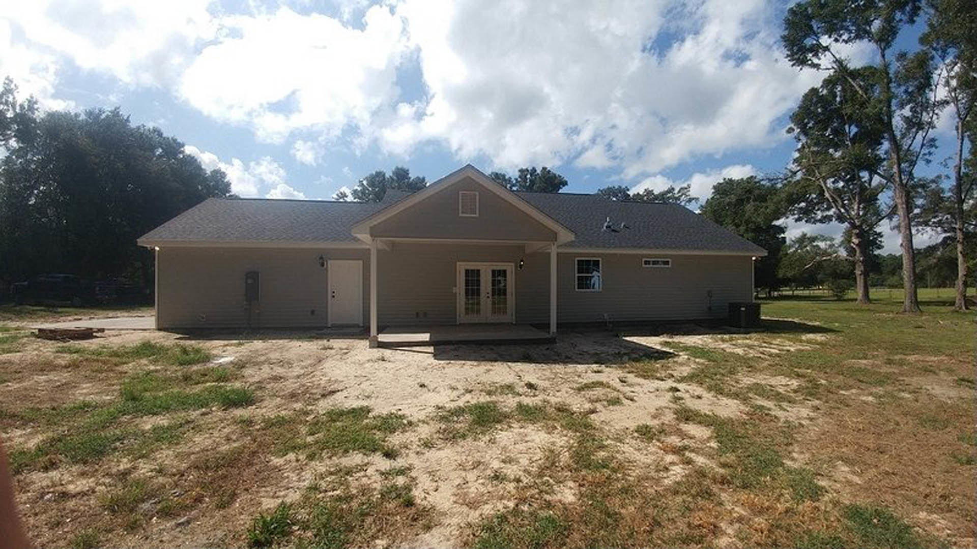 Two-story home with double glass-paneled front doors, covered porch, light siding, large grassy yard, dirt patch near entry, and mature trees in the background under a partly