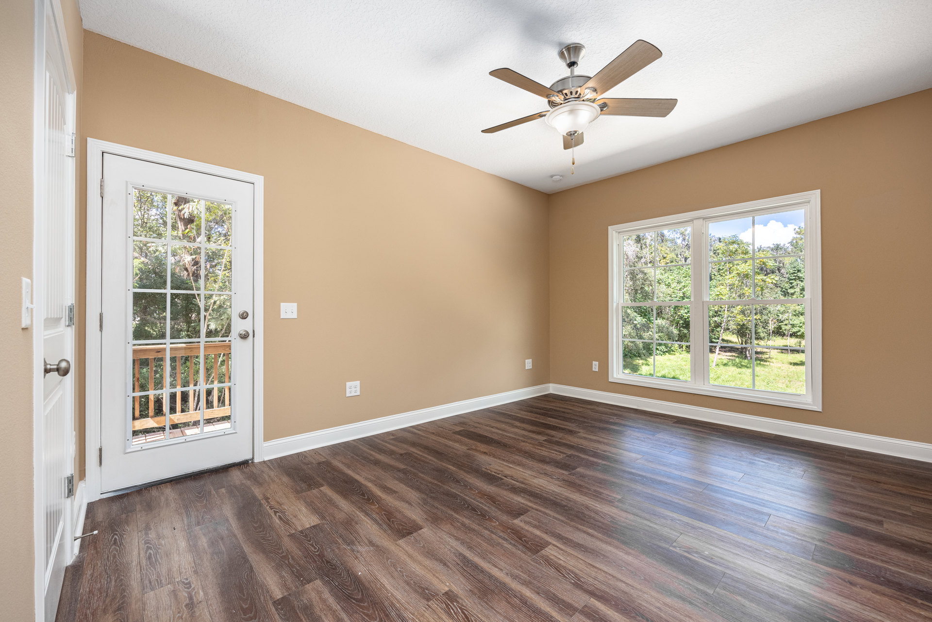 Ceiling fan with light fixture above hardwood floor, glass-paneled door and window offering views of trees, wood railing visible through window