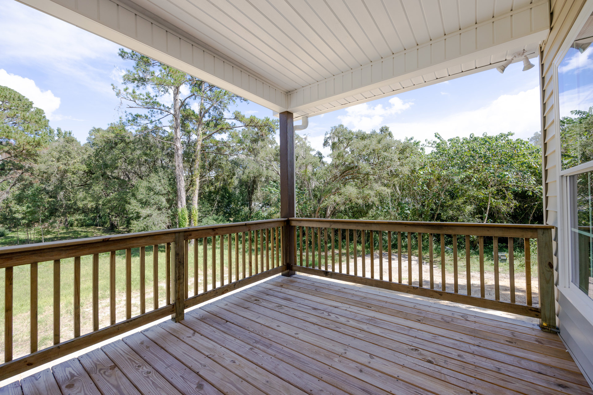 Wooden deck with metal railing overlooking leafy trees, shaded outdoor space with balusters and sky visible in background