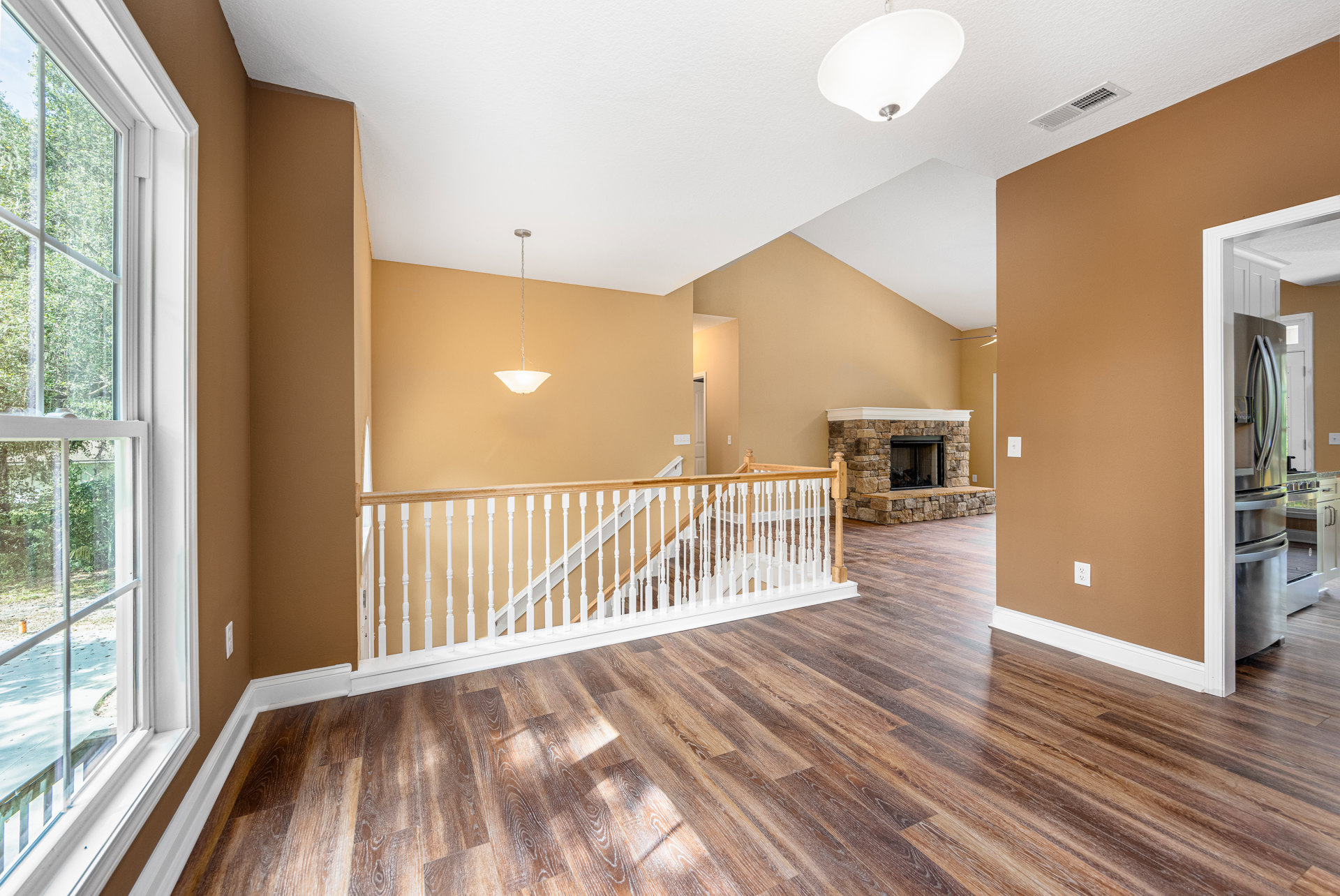 Living room with hardwood floors, white staircase railing, stone fireplace featuring a wood mantel, large window overlooking trees, white ceiling with a light fixture and hanging