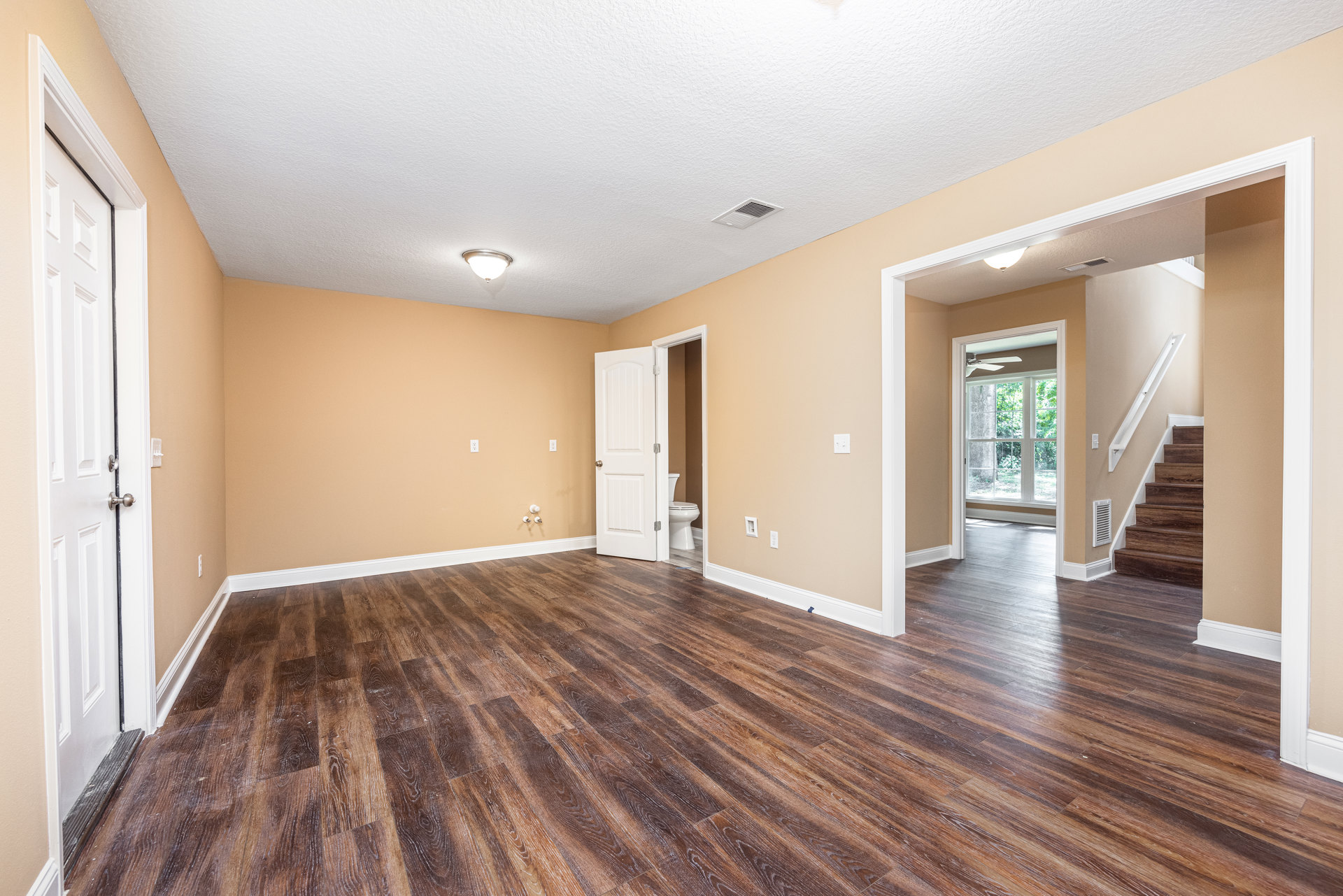 Wood flooring in a bright room with white-framed window, ceiling light fixture, staircase, and white door leading to a bathroom with visible toilet