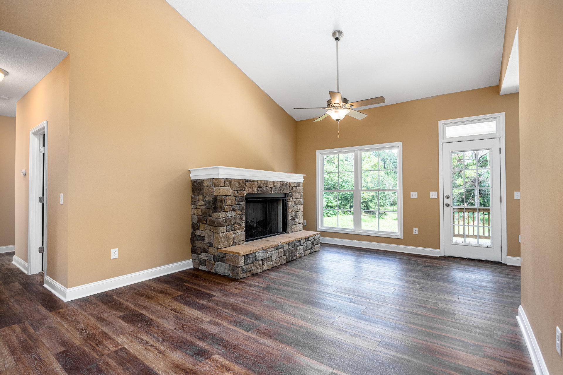 Living room with wood flooring, white-framed fireplace with screen, ceiling fan, large window showing trees, glass-paneled white door, and metal railing