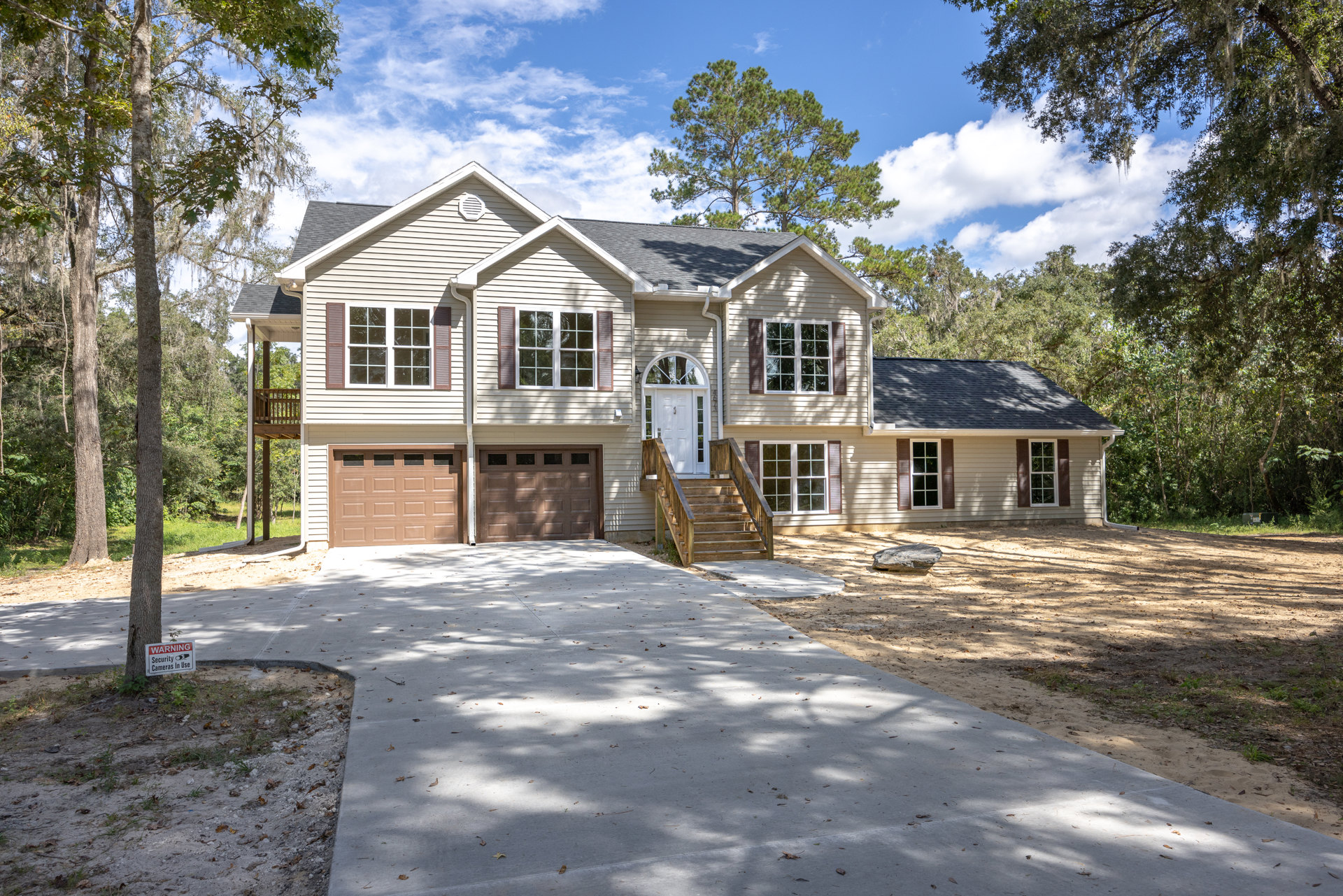 Two-story home with white-trimmed windows, wooden exterior stairs, paved driveway, mature trees, lawn, and a signpost near a large rock