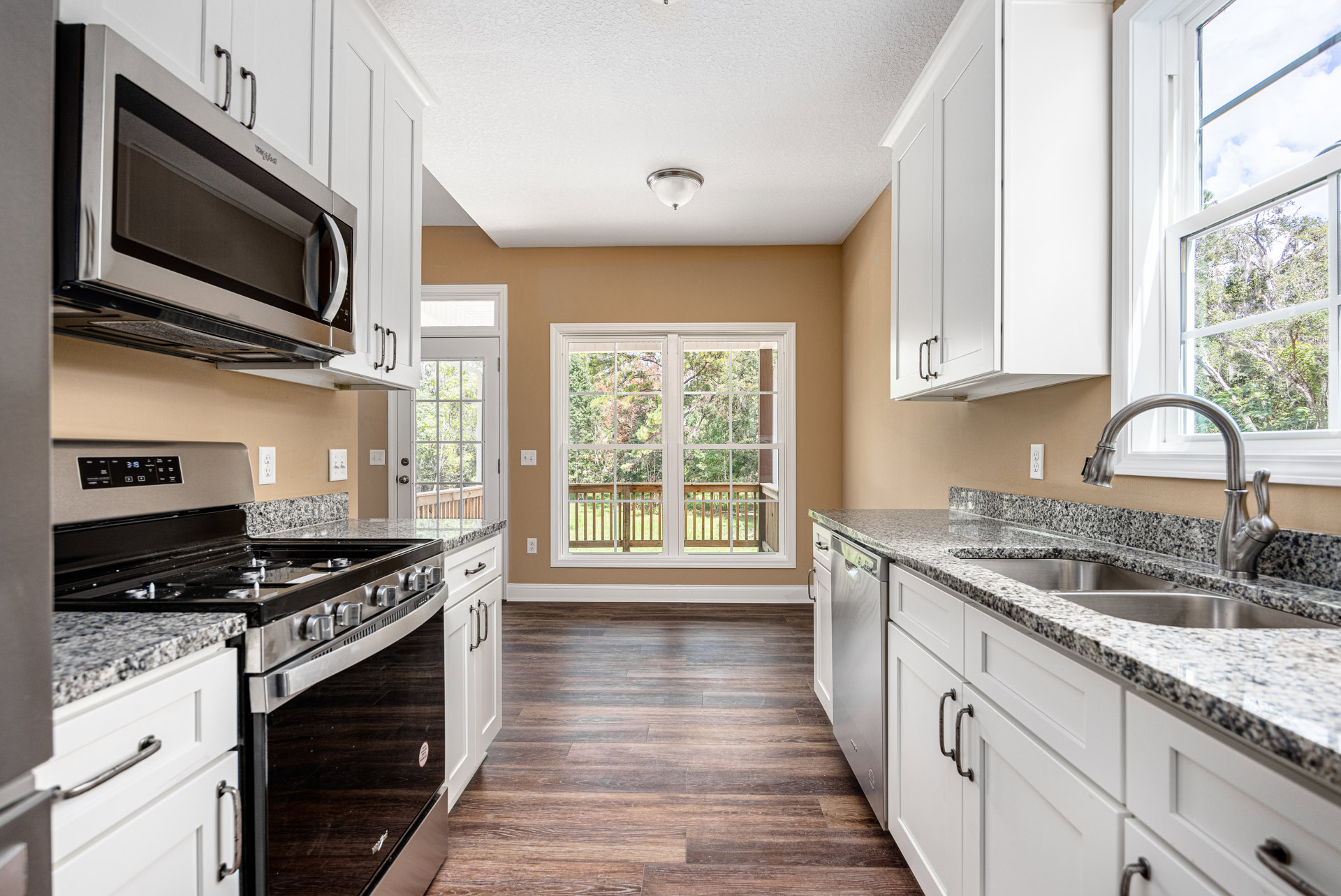 White kitchen cabinets with granite countertops, stainless steel stove and microwave, ceiling light fixture, window overlooking deck and trees.