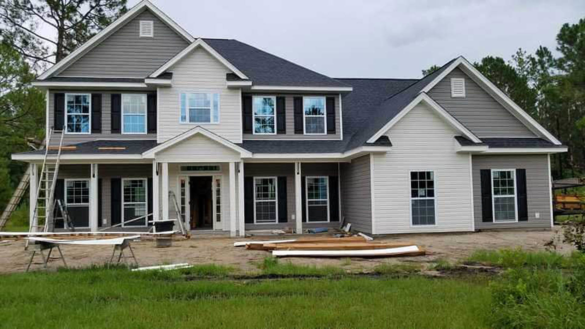 Gray-sided house under construction with white-framed window, white vent, ladder leaning against wall, and green lawn in foreground