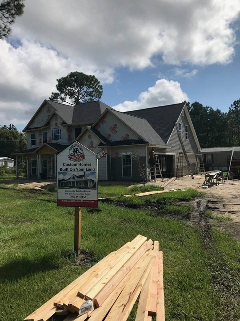 Partially built house with exposed wooden framing, pile of lumber on grassy lot, construction sign on post, large leafless tree nearby, overcast sky overhead