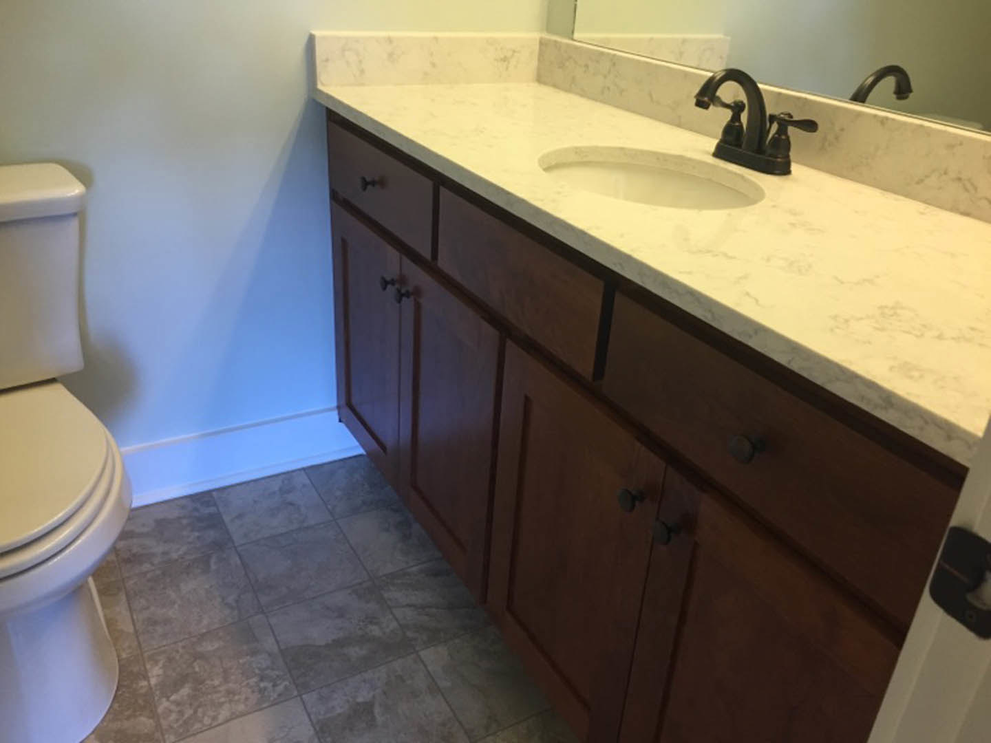 Bathroom with marble countertop, undermount sink, chrome faucet, white toilet, grey tile floor, wall-mounted mirror, and white door