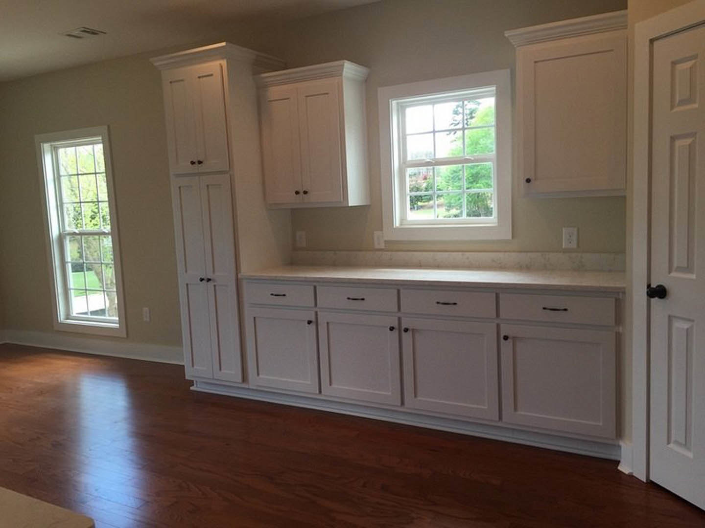 White kitchen cabinets, wood flooring, large windows with views of trees, close-up of a door handle, white cabinet doors