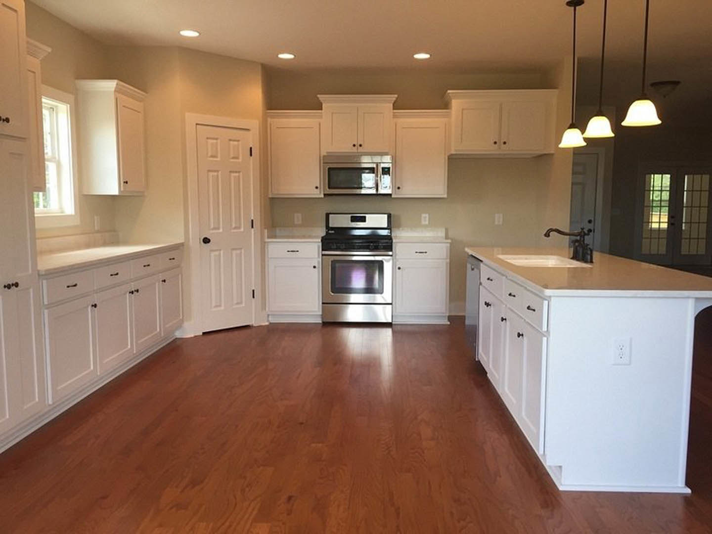 White kitchen with shaker cabinets, hardwood flooring, white island featuring an undermount sink, built-in microwave, white door with black handle, and white electrical outlet with