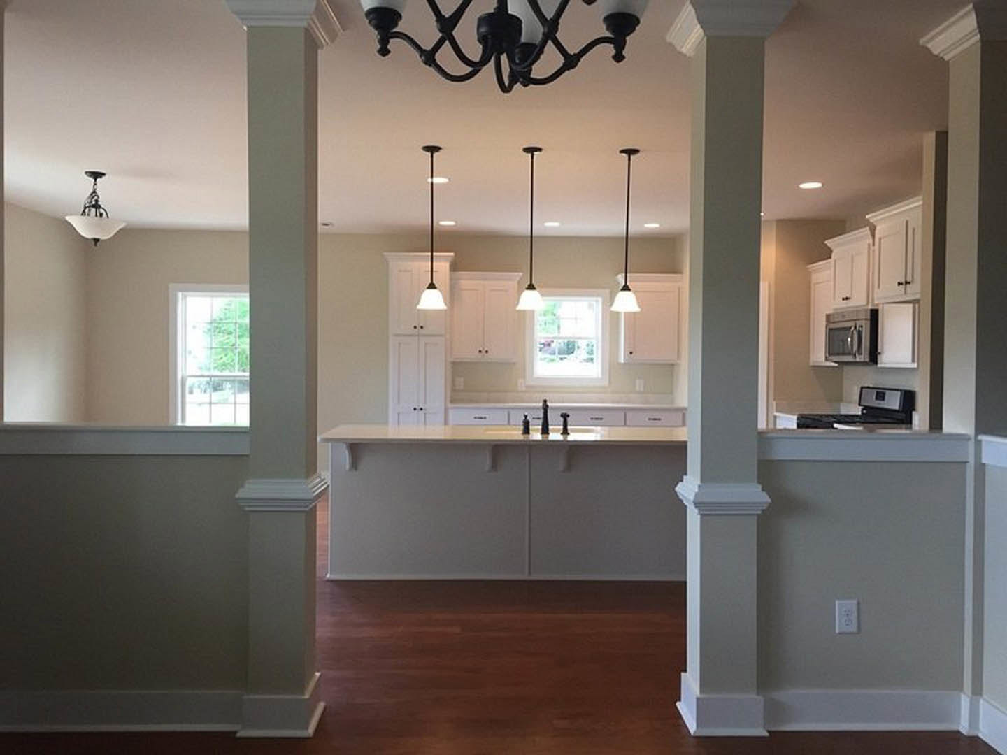 Kitchen with white pillars, a central chandelier, white cabinetry, light countertops, and a window with a white frame; visible black light fixture, microwave, ceiling details, and