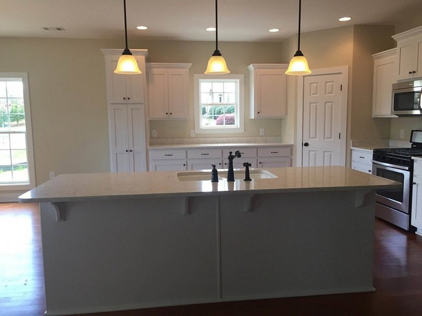 White kitchen with shaker cabinets, large central island, white countertops, black faucets, stainless steel microwave and oven, window with white frame overlooking garden.