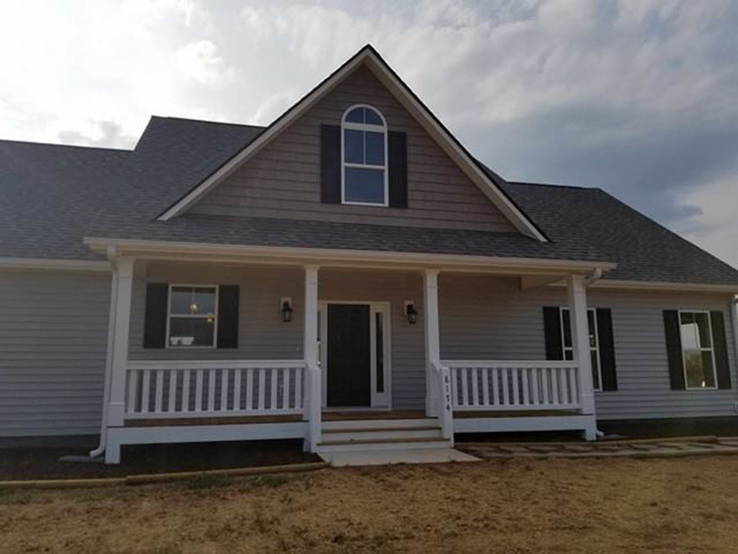Two-story home with gray siding, white trim, covered front porch featuring white railing, large windows reflecting trees, and dark front door