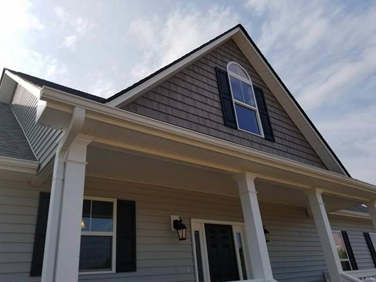Front porch with white siding, black door with white trim, sash window illuminated from inside, gabled roof against blue sky