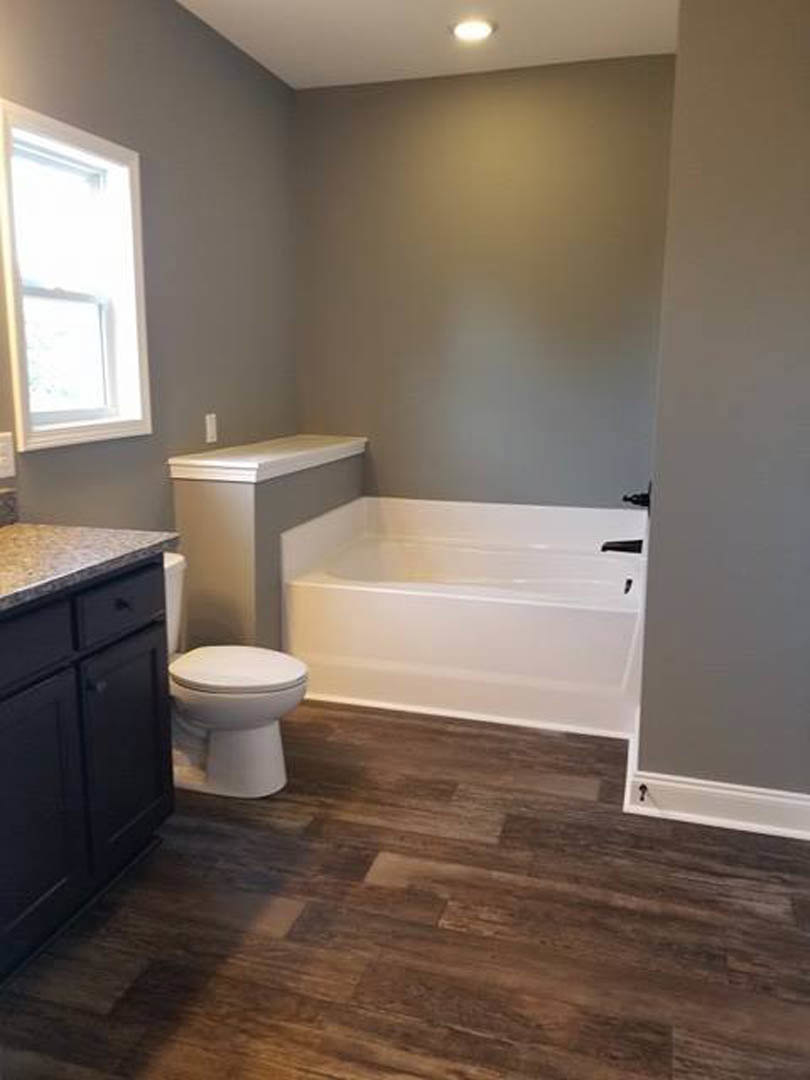 Bathroom with a white toilet beside a freestanding bathtub, black vanity cabinet with matching handle, wood flooring, and window letting in natural light
