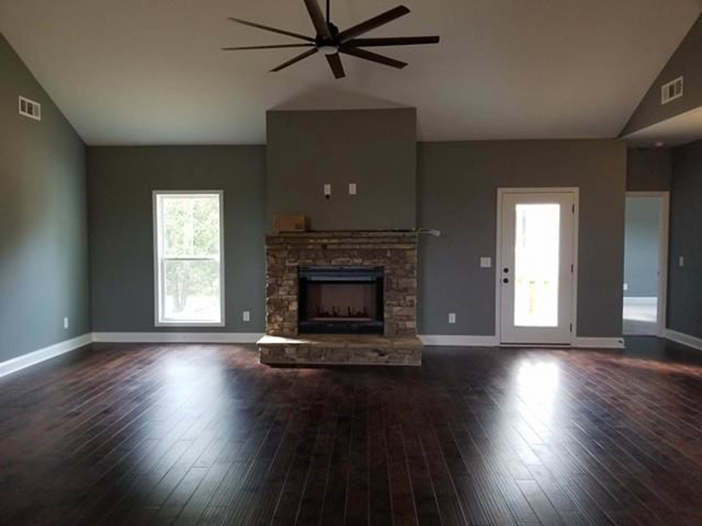 Living room with wood flooring, white ceiling and ceiling fan, black-framed fireplace, white-framed window, and white door illuminated by natural light