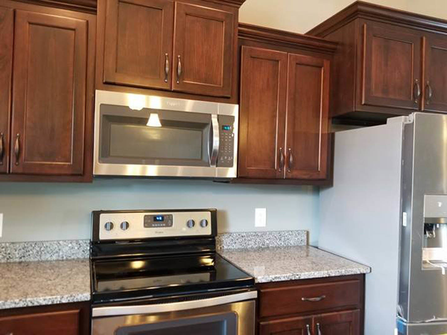 Modern kitchen with white shaker cabinets, stainless steel microwave oven mounted above a matching stove, granite countertops, and a stainless steel refrigerator