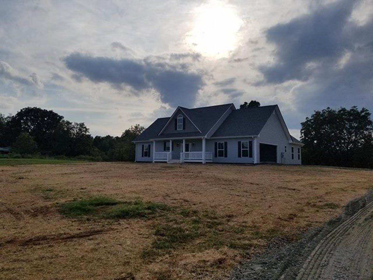 Two-story house with gray roof and white siding, front porch, square windows, surrounded by a large grassy lawn and leafy trees under a partly cloudy sky
