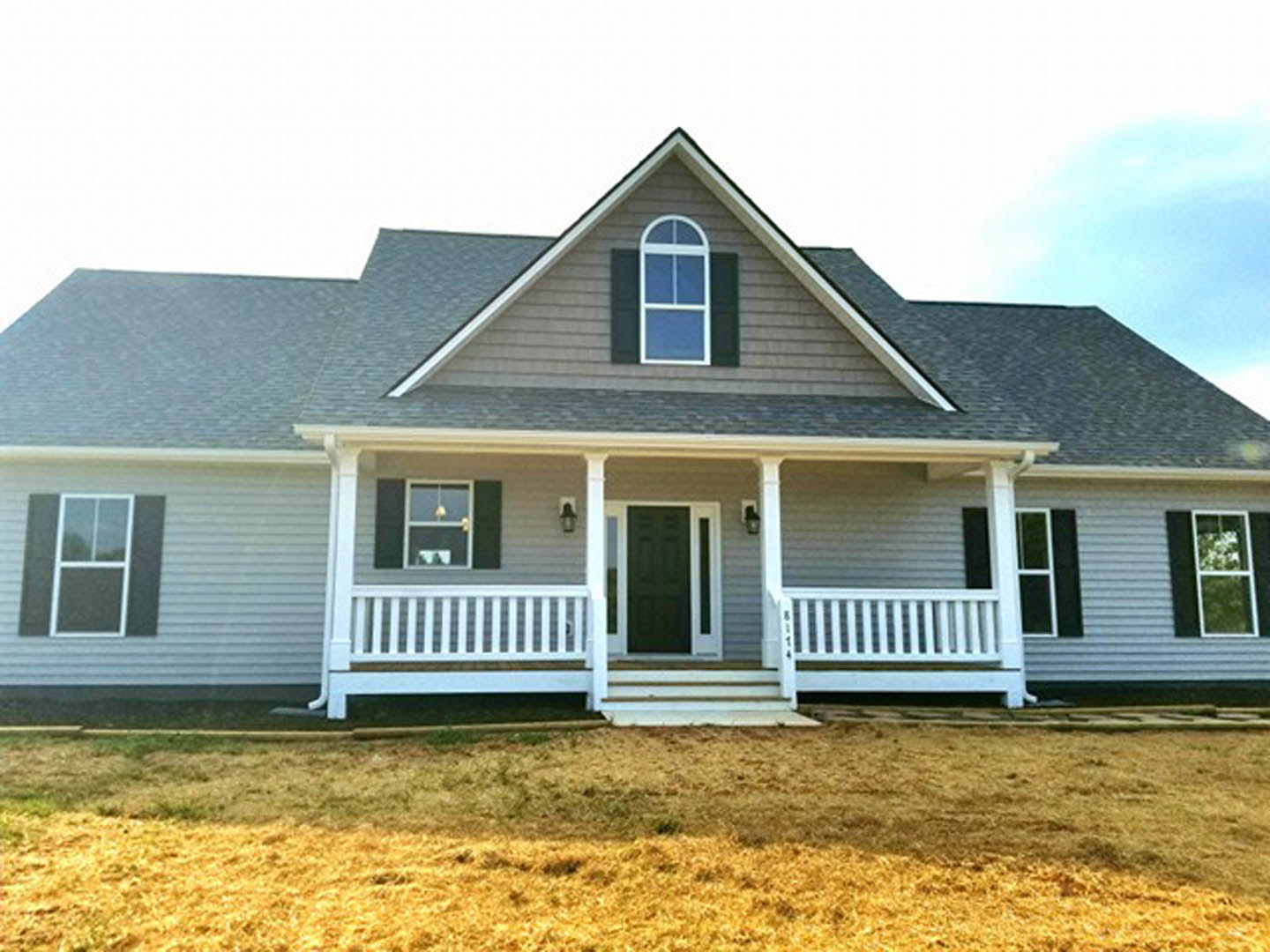White farmhouse with black front door, wide porch featuring white railing with black accent stripe, manicured green lawn, large windows, and a white bench.