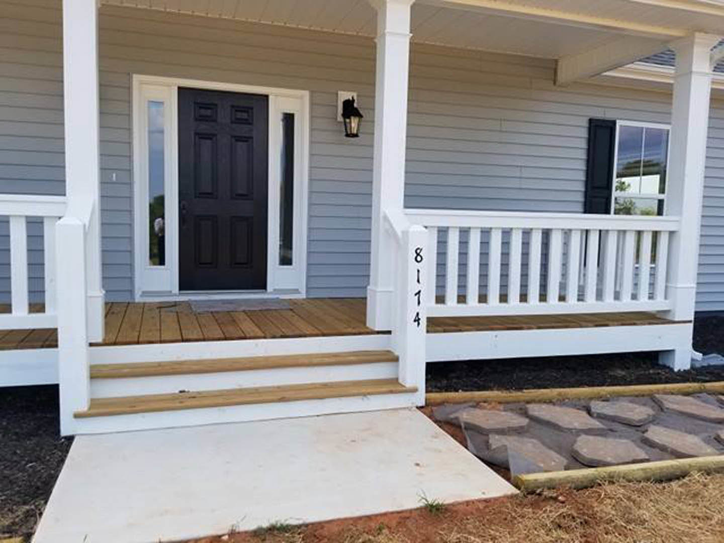 White siding house with black front door, white porch railing, concrete steps, and stone pathway; partial view of a person’s head in foreground.