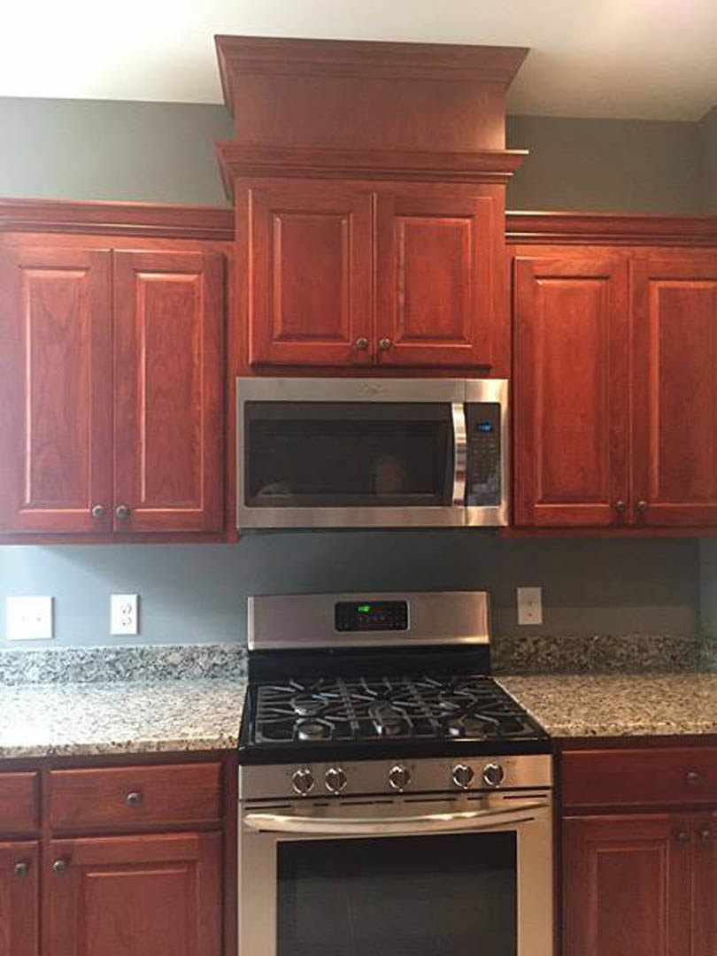 Modern kitchen with stainless steel stove and oven, microwave mounted above, white cabinetry, stone countertop, and a window reflecting a person's head.