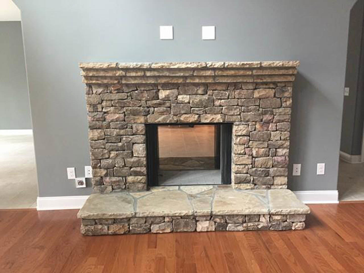 Stone fireplace with glass door and fire screen set in a textured stone wall, wood floor with stone ledge, window to the side, white square accent on gray surface.