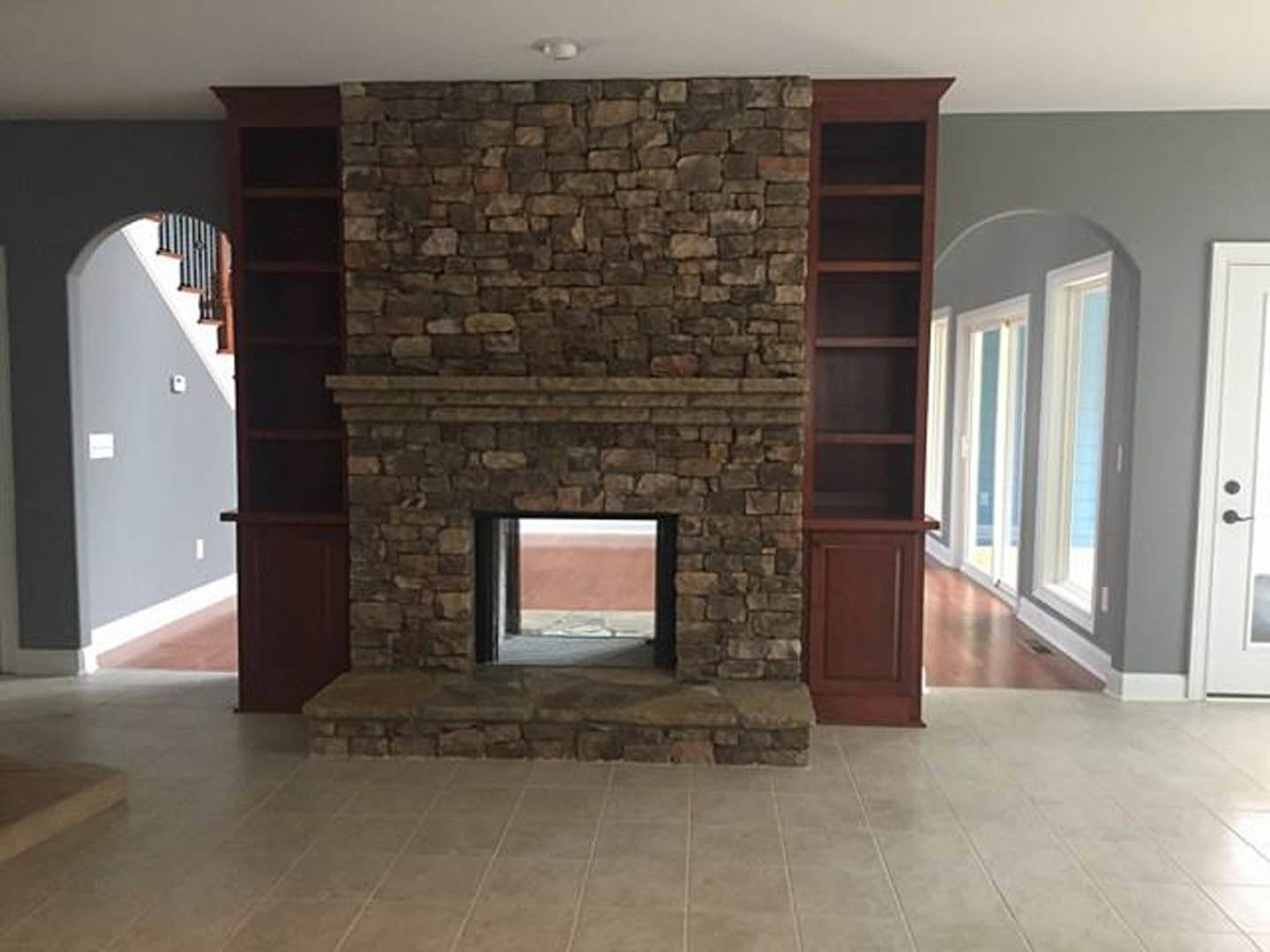 Stone fireplace with white mantel, built-in shelves, large window framed by brick accent wall, hardwood floors, and white walls with light switch.
