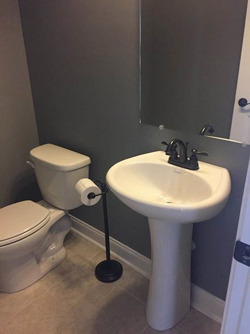 Bathroom with white pedestal sink, white toilet with closed lid, wall-mounted toilet paper holder, light-colored tile floor, and neutral painted walls.