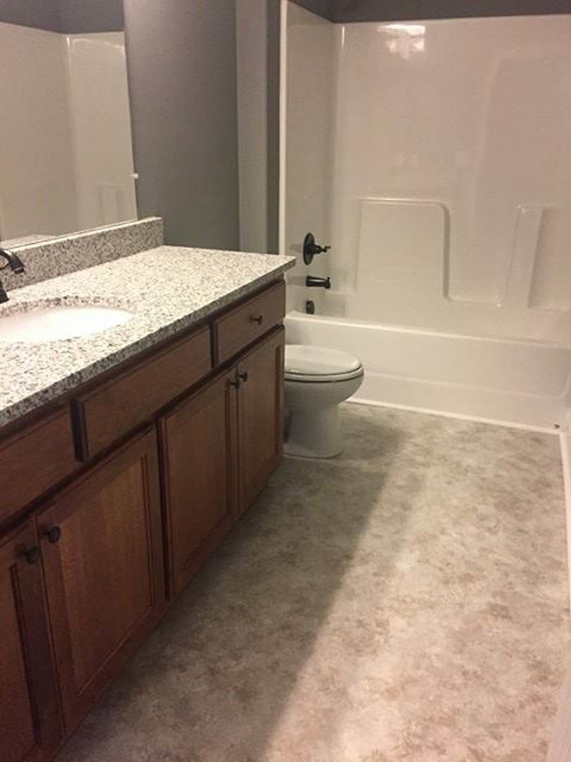 Bathroom with granite countertop, undermount sink, chrome faucet, white toilet, glass-enclosed shower, light tile flooring, and white cabinetry