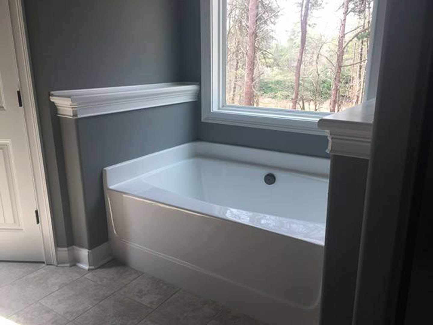 Freestanding white bathtub with chrome faucet beside large window, gray tile floor, and white walls in modern bathroom