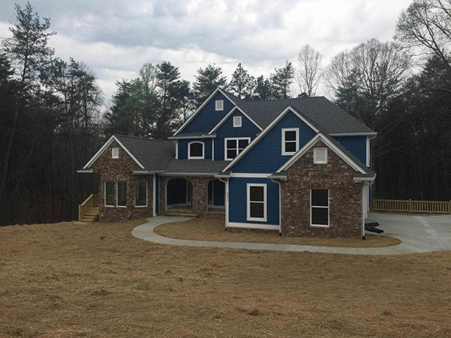 Two-story blue house with brick and stone facade, white-trimmed windows, paved driveway, mature trees, and cloudy sky