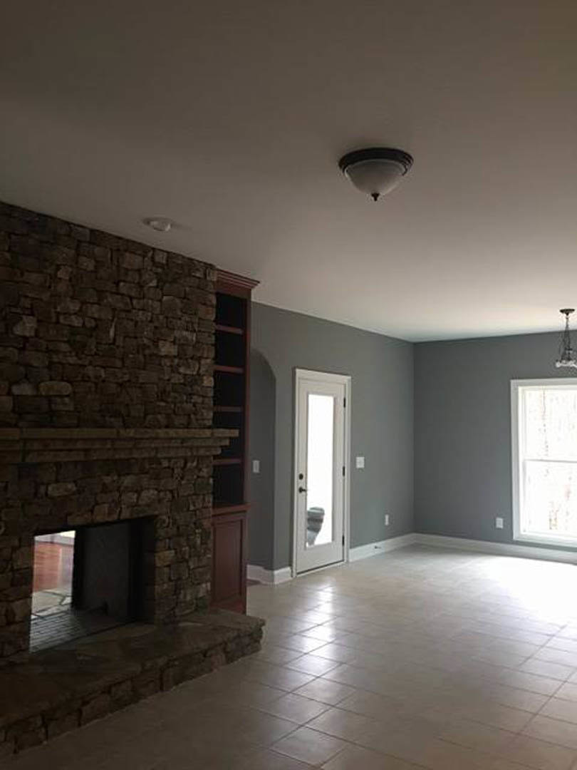Brick fireplace with white mantel, adjacent white door letting in natural light, neutral tiled floor, pale walls, and ceiling visible in custom home living room.