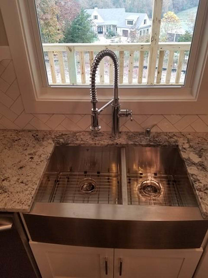 White kitchen sink with chrome faucet set in a stone countertop beneath a large window, light streaming onto tiled backsplash and neutral walls.