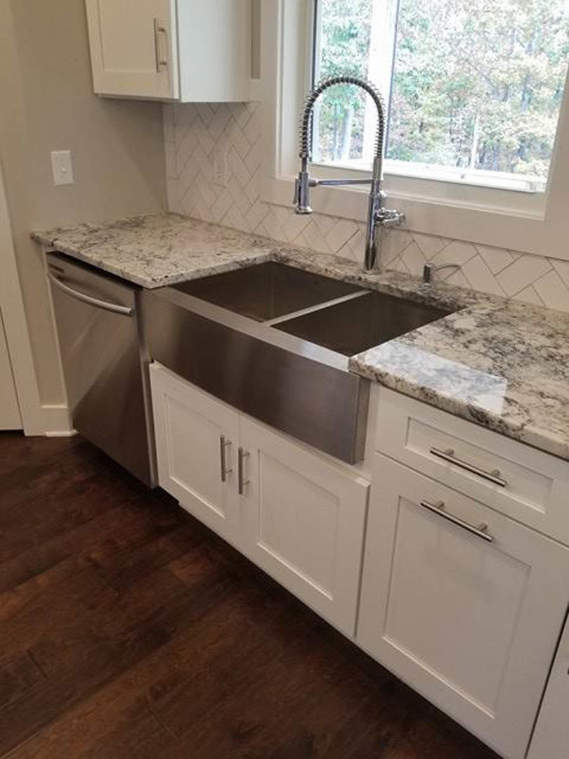 Modern kitchen featuring white cabinetry with silver handles, stainless steel sink beneath a window, tiled backsplash, stone countertop, and stainless steel refrigerator