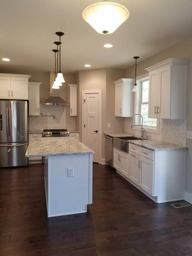 White kitchen with shaker cabinets, stainless steel refrigerator, marble island countertop, black hardware, pendant light fixture, and hardwood flooring.
