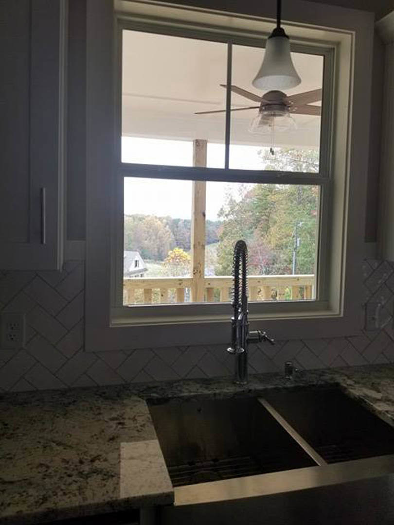 White kitchen sink under a large window, chrome faucet, marble countertop, white tile backsplash, natural light illuminating the space