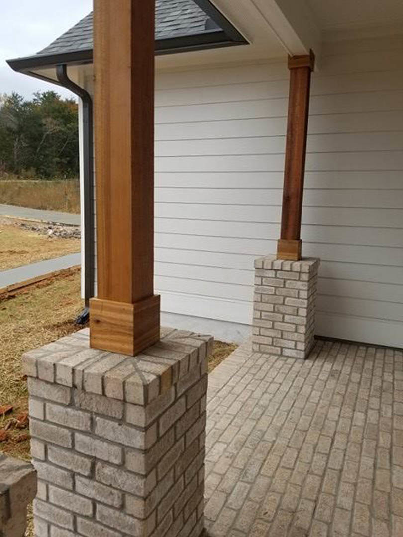 Red brick pillar supporting wooden post beside white door, brick flooring, exterior of custom home