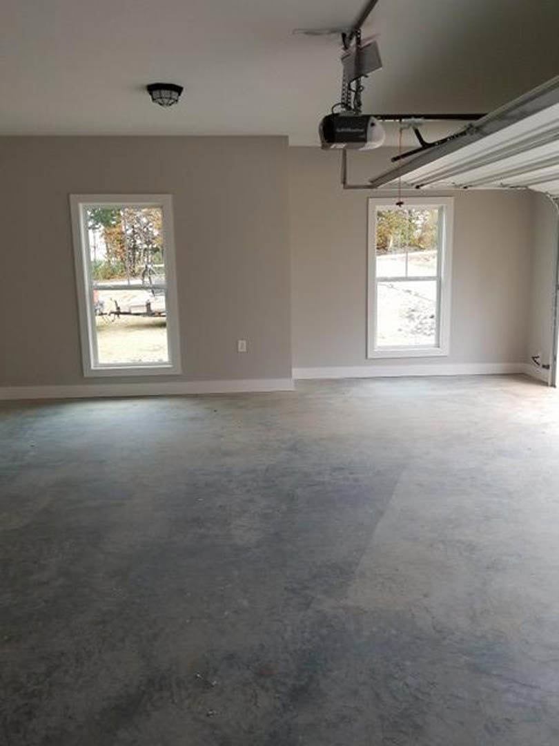 Garage interior with concrete floor, plaster walls, large windows providing daylight, ceiling fan, and overhead light fixture