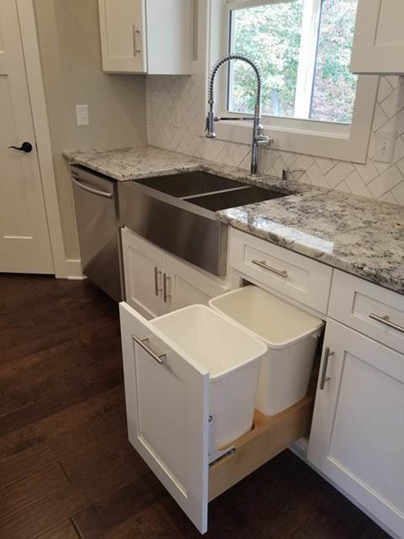 Stainless steel sink with chrome faucet set in a stone countertop, wood flooring, white trash can in pull-out cabinet drawer, tile backsplash, and white cabinetry